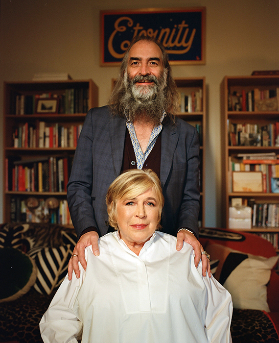 Marianne Faithfull, a white woman with short blonde hair wearing a white blouse, sits in front of Warren Ellis, a tall man with a long grey beard. Both are smiling. In the background, there are shelves full of books.