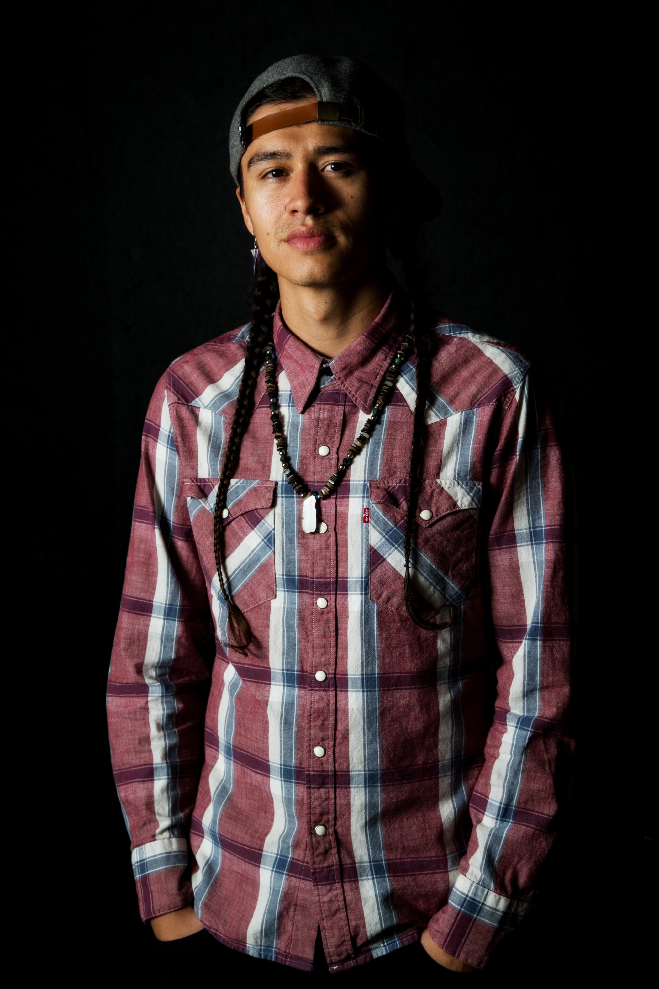 A young man with braids and a backwards baseball cap stands against a black backdrop wearing a plaid Levi's button-down shirt.