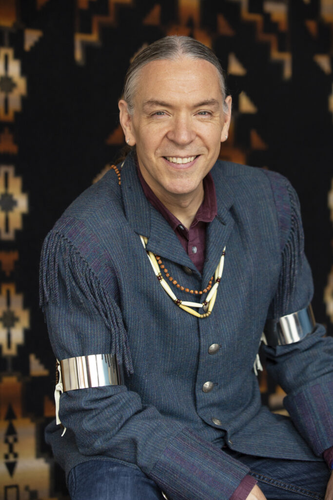 A middle-aged man dressed in a denim shirt, traditional arm braces and necklaces sits facing the camera smiling in a head shot.