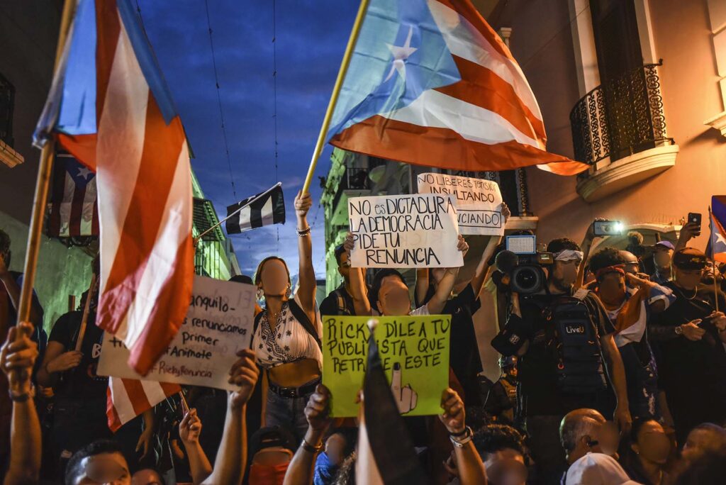 A group of protesters in a street hold Puerto Rican flags and bear signs criticizing the Puerto Rican governor.
