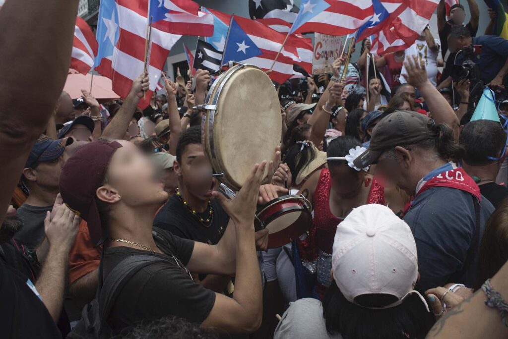 A close-up photo of people playing hand drums amid a crowd waving the Puerto Rican flag.