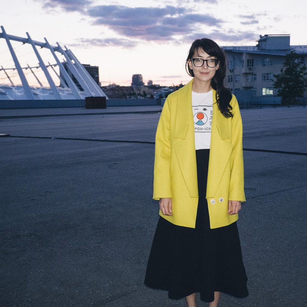 A woman wearing black-rimmed glasses and stylish yellow coat stands in the foreground of an urban horizon