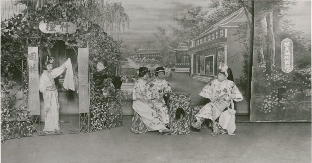 Three actors sit around a small table at the center of a stage. On the left, a woman stands in a doorway while a figure dressed in black peers at her.
