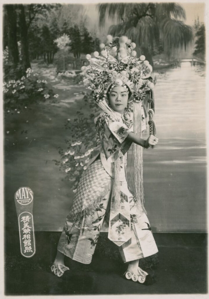 A performer wearing a headdress poses in a braced position, her right arm straightened and projected across the front of her body.