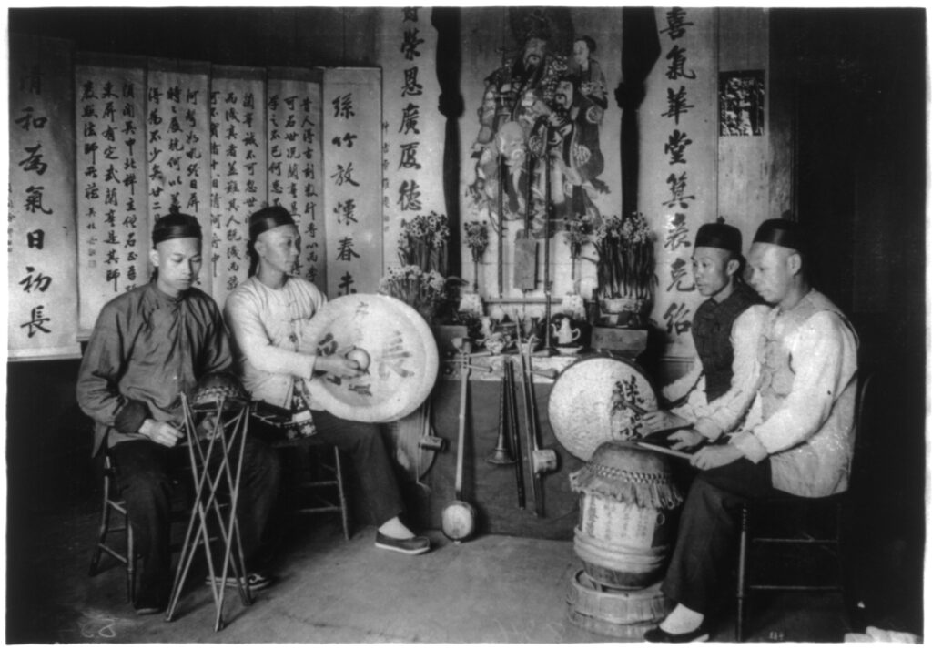 Four Chinese musicians sit in a semi circle with other instruments leaning against the wall, which displays calligraphy.