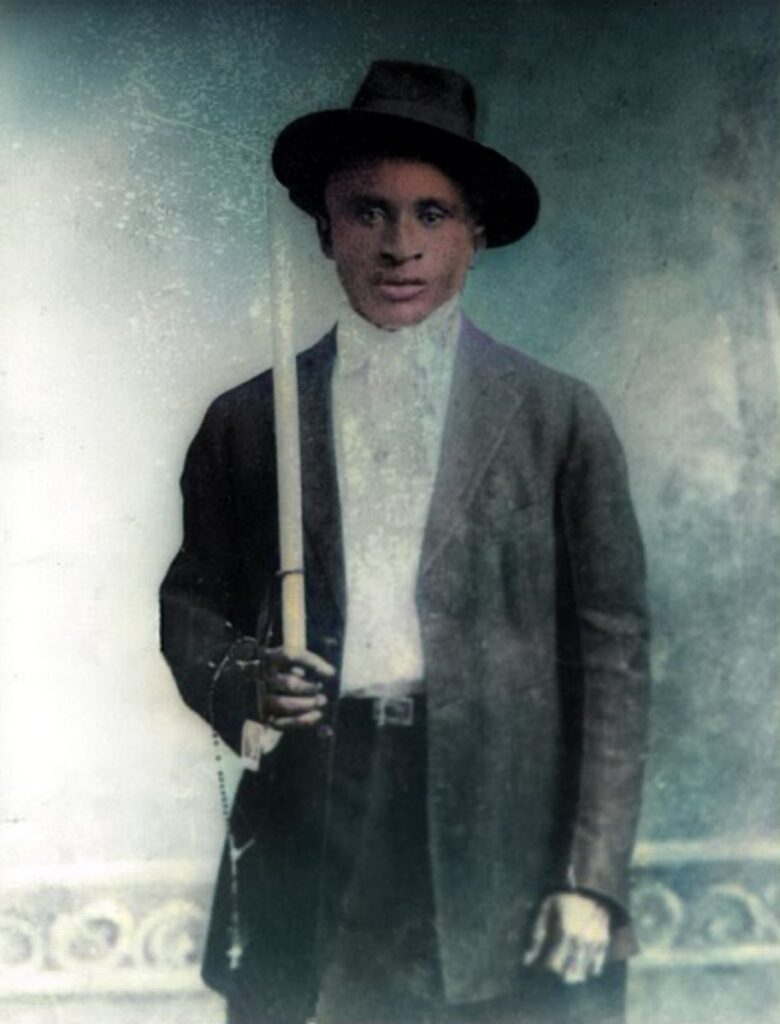 A Louisiana Creole teenage boy circa 1912 looks directly at the viewer wearing a bowed white shirt and dark suit and hat while holding a rosary and tall white confirmation candle.